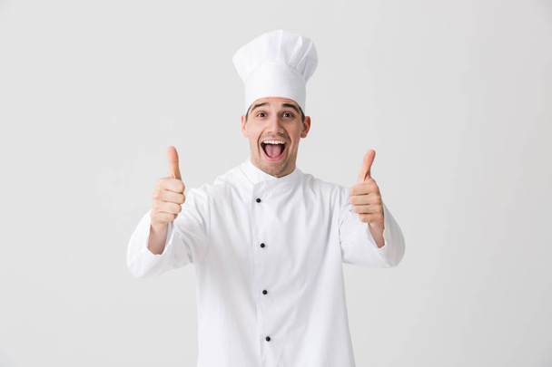 Зображення красивого збудженого молодого кухаря в приміщенні ізольовано на білому тлі стіни, показуючи великі пальці вгору жест
. - Фото, зображення