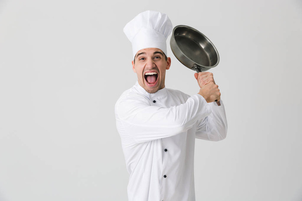 Image of excited emotional young man chef indoors isolated over white wall background holding frying pan. - Φωτογραφία, εικόνα