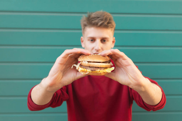 Nuori mies seisoo turkoosi tausta ja pitää herkullinen hampurilainen käsissään. Pikaruokakäsite
 - Valokuva, kuva