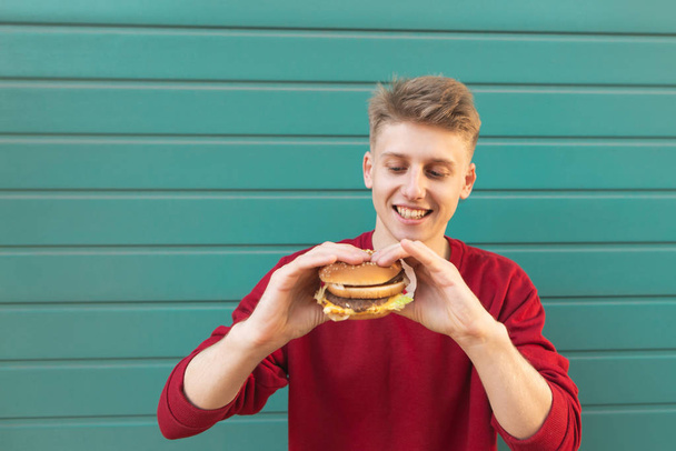Hongerige man met voedsel in zijn handen tegen de achtergrond van een groene muur. Portret van een knappe jonge man met een hamburger in zijn handen, kijken naar een sandwich en lachend op een turkooizen achtergrond. - Foto, afbeelding