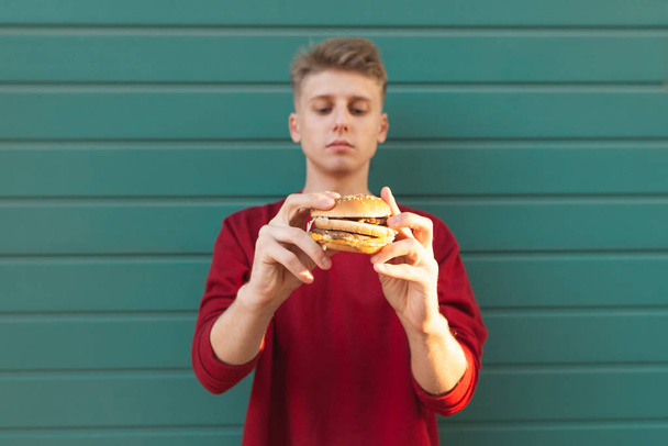 美しい若い男はターコイズ色の壁の背景の上に立って、食欲をそそるバーガーを保持します。ハンバーガーに焦点を当てます。ハンバーガーは、学生の手で. - 写真・画像