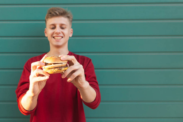 色の壁の背景に彼の手でハンバーガーと立っている若い男の笑みを浮かべてします。ファーストフード。彼の手でハンバーガーに若いティーネー ジャー。ハンバーガーに焦点を当てる - 写真・画像