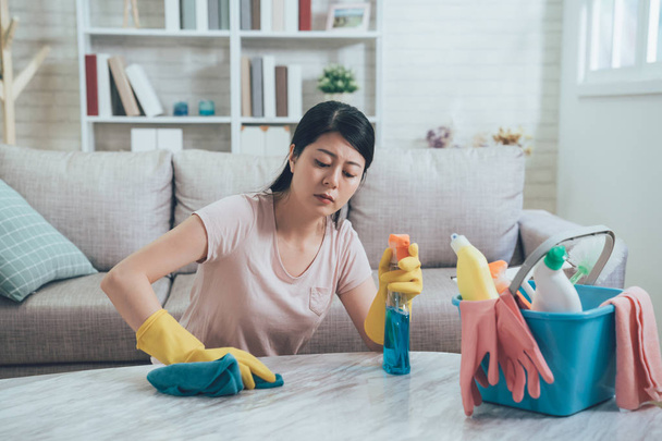 Pretty asian woman doing her house chores in cozy sunlight living room at home. young housewife wiping table by blue rag and protective gloves in hands. lady sitting on floor in front of couch. - Foto, imagen