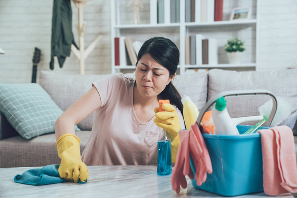 asian housewife wearing gloves wiping marble table with all of her power by rag. young lady holding spray and trying to remove the dust. frowning female doing housekeeping in apartment. - Foto, afbeelding