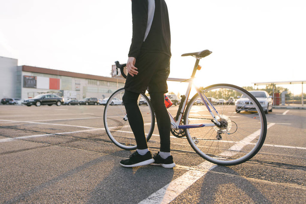 Велосипедист в тёмной спортивной одежде с велосипедом на фоне парковки на закате. Спортивная концепция. Экологический транспорт
. - Фото, изображение