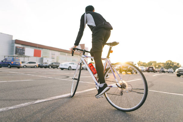 Takana pyöräilijä ratsastaa valkoinen valtatie pyörä parkkipaikalla, auringonlasku ja moderni arkkitehtuuri taustalla. Ihmiset ja liikenne. Harrastuspyöräily.Copyspace
 - Valokuva, kuva