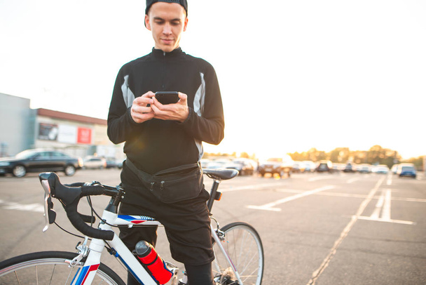 Αστεία νεαρός άνδρας με ένα ποδήλατο στέκεται ενάντια σε το φόντο του ηλιοβασιλέματος, κοιτάζοντας την οθόνη του smartphone και να χαμογελά. Ευτυχισμένος ποδηλάτης χρησιμοποιεί ένα smartphone. - Φωτογραφία, εικόνα