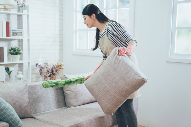 asian wife using green feather duster carefully cleaning the sofa. young female housekeeper taking up pillow dusting the couch in the cozy living room at apartment. lady doing house chores in apron. - Photo, Image