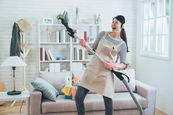 cheerful asian wife wearing earphones playing vacuum cleaner as a guitar enjoy music dancing while doing housework in the living room. young housewife joyfully doing house chores at home. - Фото, изображение