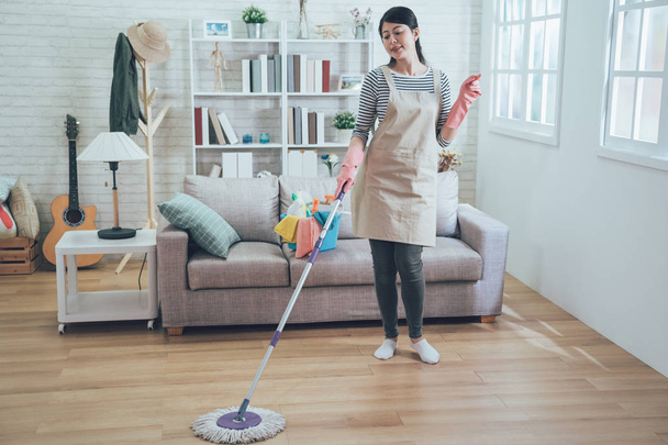female housekeeper wearing apron and pink rubber gloves mopping the floor. young asian housewife singing clicking her finger relaxing doing housework cleaning in living room in daytime cozy apartment - Photo, image