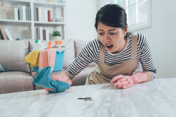 asian housewife with a shock frowning face cleaning the marble table with dirt on it. lady unsatisfied opening mouth doing housework at home. woman in apron and protective gloves using rag wiping. - 写真・画像