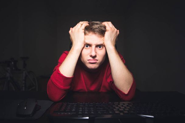Retrato de un jugador frustrado mirando un monitor de computadora con una expresión de caras tristes. Joven se sienta a la mesa en la noche en casa y juega juegos en un ordenador
. - Foto, Imagen