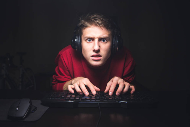 Retrato de jugador enfocado mirando el juego y usando un teclado de computadora. Un joven concentrado en los auriculares juega juegos en línea en casa en una computadora y se centra en mirar a la cámara
. - Foto, imagen