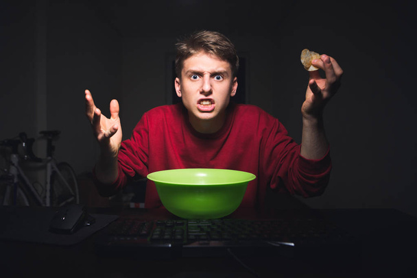 Młody chłopak z frytkami w ręku patrzy się na ekran komputera z oburzenia. Nastolatek jest patrząc ze złością na monitor podczas oglądania filmów w Internecie i jedzenia przekąski. - Zdjęcie, obraz