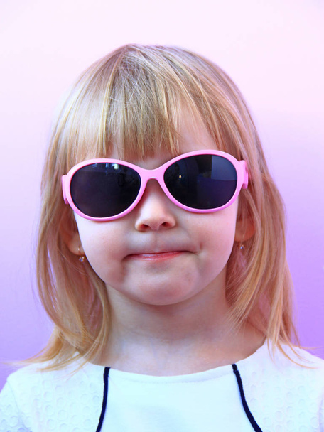 Портрет маленькой девочки в солнечных очках на розовом фоне. Студийный снимок молодой девушки в розовых солнечных очках. Модный маленький ребенок в солнечных очках. Студийный портрет очаровательной малышки
 - Фото, изображение