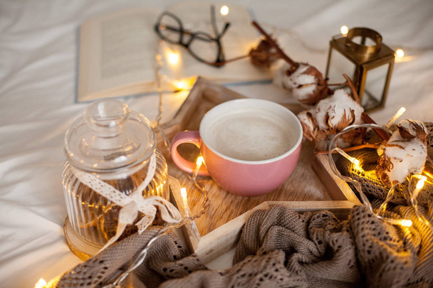 Mok warme cappuccino op een houten dienblad is op het bed. Gezellige inrichting. Ontbijt. Mok, plaid, katoen, kaars. Boek. Kerstverlichting. Vakantie. - Foto, afbeelding