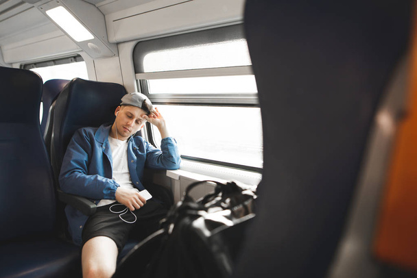Joven durmiendo en un tren cerca de la ventana con auriculares y un teléfono inteligente en sus manos. El somnoliento pasa junto al tren por la ventana
. - Foto, imagen