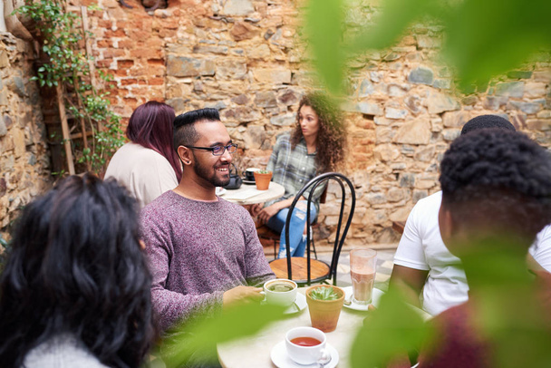 Joven sonriente hablando con amigos sobre el café mientras se sientan juntos alrededor de una mesa en el patio de un café de moda
 - Foto, Imagen