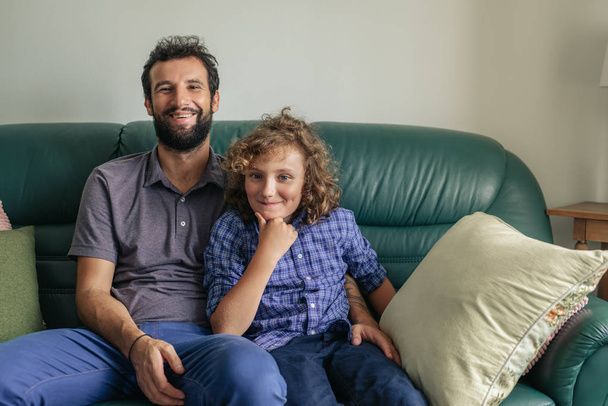 Portrait of a smiling single father and his cute young son sitting together on their living room sofa at home - Photo, Image
