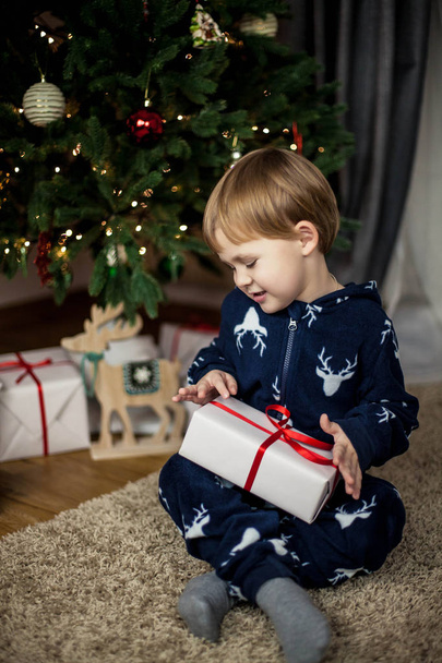 Pieni söpö poika istuu lähellä joulukuusi ja tilalla laatikko lahja. Juhlapyhät. Joulu. Uusi vuosi. 3-vuotias lapsi joulupyjamassa
. - Valokuva, kuva