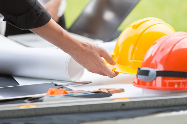 Close-up van mannelijke ingenieur harde staande op de werkplek. Helm met huis model en oranje dak, werken met de Tablet PC- en computer- en witte plan - Foto, afbeelding