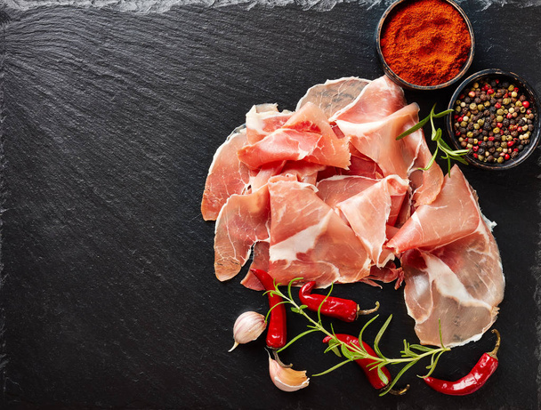 spanish dry-cured ham, jamon curado thinly sliced and served on a black stone plate with rosemary and species, horizontal view from above, flat lay, copy space - Zdjęcie, obraz