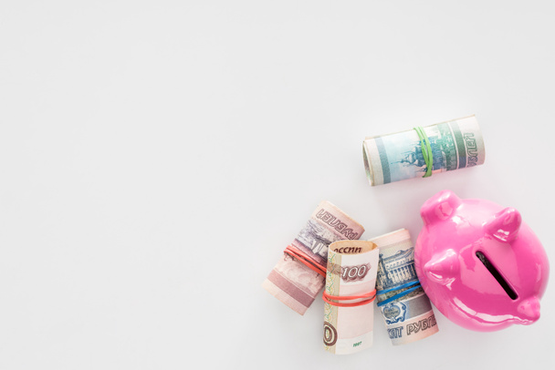 Draufsicht auf rosa Sparschwein und verschiedene gerollte russische Banknoten auf weißer Oberfläche - Foto, Bild