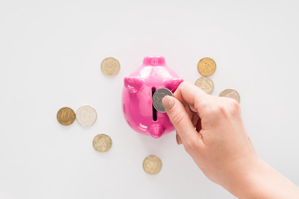 περικομμένη εικόνα της γυναίκας βάζοντας κέρμα σε ροζ κουμπαράς σε λευκή επιφάνεια - Φωτογραφία, εικόνα