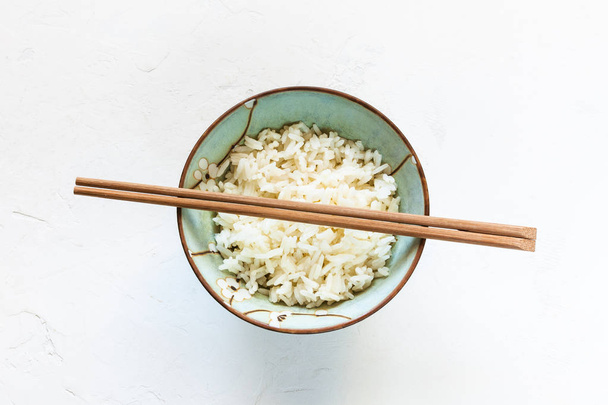 Κάτοψη των ξύλινων chopsticks πάνω από το φλιτζάνι με το βρασμένο ρύζι στο λευκό του σκάφους σκυροδέματος - Φωτογραφία, εικόνα