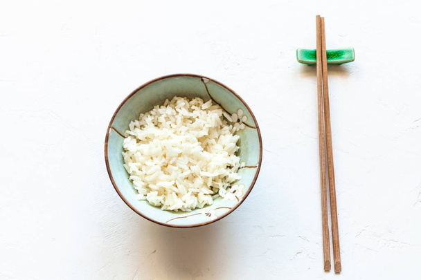 κάτοψη του φλιτζάνι με βρασμένο ρύζι και ξύλινων chopsticks για υπόλοιπο στο λευκό του σκάφους σκυροδέματος - Φωτογραφία, εικόνα