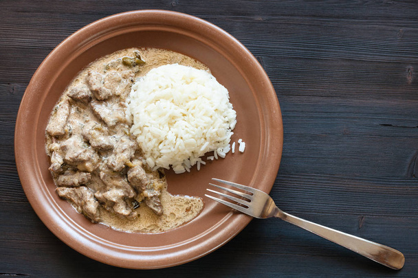Russian cuisine dish - prepared for eating a portion of Beef Stroganoff (Beef Stroganov, Befstroganov) pieces of stewed meat in sour cream with boiled rice on brown plate on dark table - Photo, Image