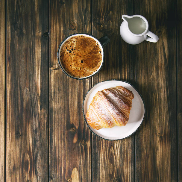 Homemade croissant with sugar powder, cup of coffee, jug of milk over wooden plank background. Flat lay, space. Square image - Φωτογραφία, εικόνα