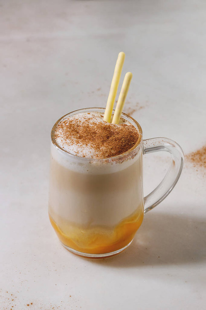 Glass of pumpkin layered spice latte with pumpkin puree, milk foam, sweet glazed sticks and cinnamon standing on white marble table. Copy space - Фото, изображение