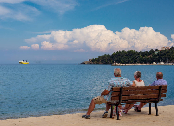 Wczasowicze turyści na ławce po prawej miejscowości turystycznej greckiej wyspy Kefalonia w Grecji na wybrzeżu Morza Jońskiego w oczekiwaniu na przybycie promu - Zdjęcie, obraz