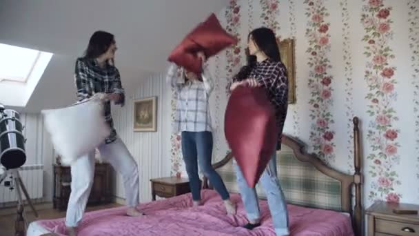 Three young pretty girls jumping on bed and fight pillows having fun at home - Záběry, video