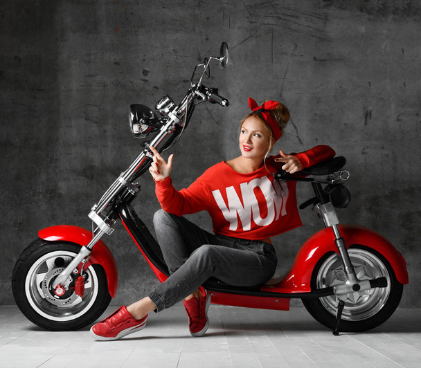Woman sitting on motorcycle bicycle scooter  retro pinup style pointing finger at the corner in red blouse and jeans - Фото, изображение