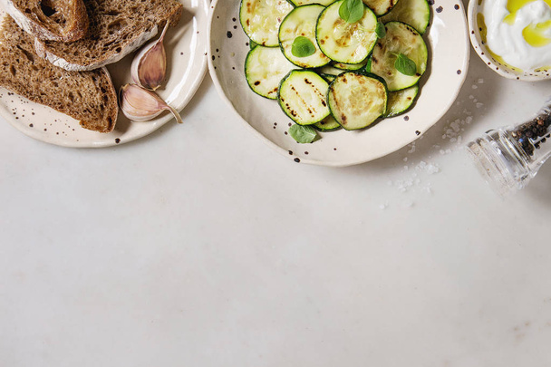 Grilled zucchini salad with yogurt dip and rye sliced bread in spotted ceramic plates over white marble background. Vegetarian food. Flat lay, space - Φωτογραφία, εικόνα