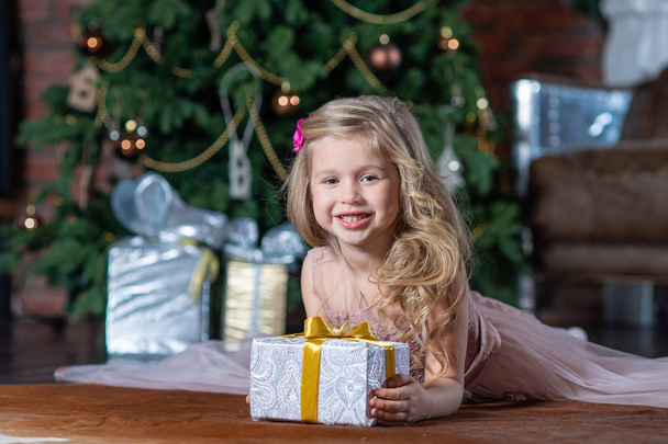 Happy little smiling girl with Christmas gift box lying on the floor on Christmas tree background - Foto, Imagem