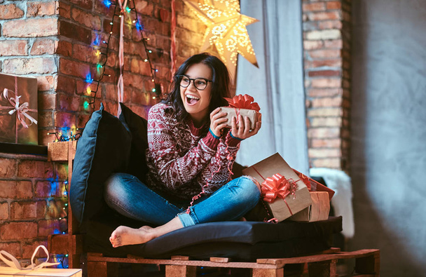 Bella ragazza godendo mattina di Natale in possesso di una scatola regalo mentre seduto su un divano in una stanza decorata con interni soppalco
. - Foto, immagini