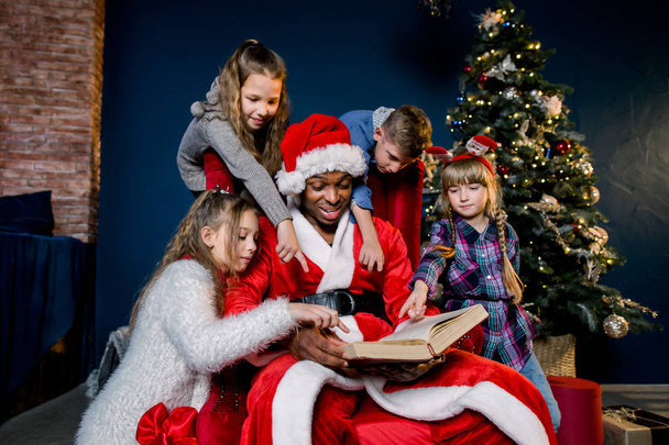 Small beautiful children listen carefully to the story Santa Claus reads while sitting on a red chair on the background of a Christmas tree. - Foto, Imagem