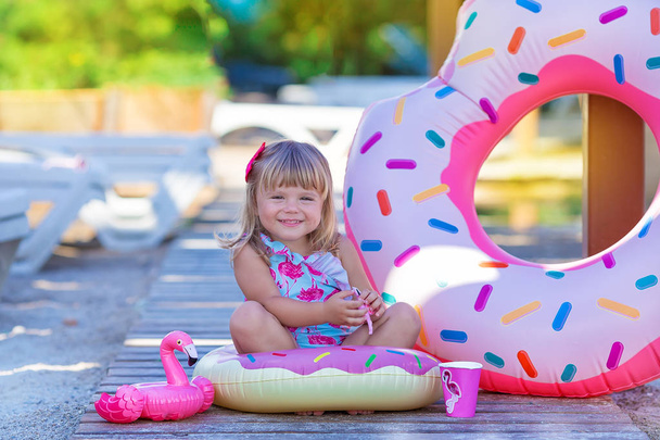Baby girl on beach with pink flamingo dressed in stylish swimming wear posing on wooden floor. Scene on vacation on sea side of a cute lady smiling and enjoy life time childhood infancy - Fotoğraf, Görsel