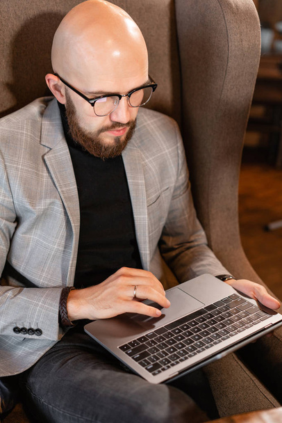 Портрет молодого человека с бородой в очках, смотрящего на монитор ноутбука. Успешные люди, бизнесмен в уютном кафе. Чтение текста во время работы над нетбуком
 - Фото, изображение
