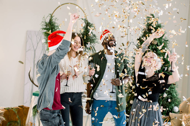 Group of four cheerful fashion dressed friends standing in front of Christmas decorations and celebrating New Year, drinking champagne - Фото, изображение