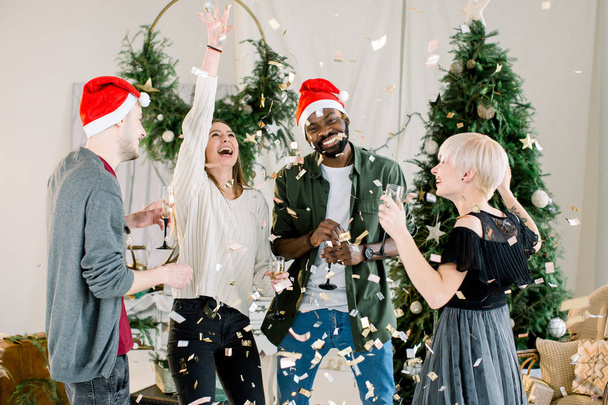 Young people in hipster outfits laughing and looking at falling golden confetti while standing in front of Christmas tree and holding glasses of champagne - Photo, Image