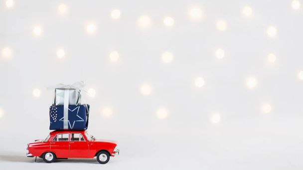 Czerwony samochodzik z prezentami świątecznymi na dachu, girlanda bokeh na tle, płytka głębia ostrości. - Zdjęcie, obraz