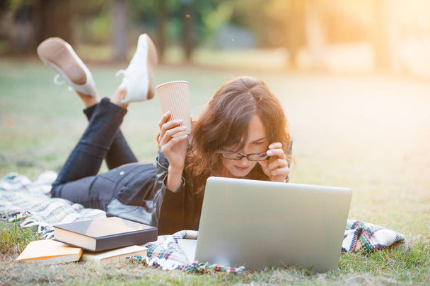Female student is studying outdoor in a park, lying down on a blanket and looking into her laptop computer with some books nearby - Photo, image