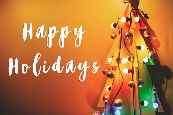 Happy Holidays texto na árvore de Natal em forma de madeira criativa com ornamentos e lâmpadas retro na parede amarela. Cartão de boas-vindas da época. Feliz Natal e Feliz Ano Novo
 - Foto, Imagem