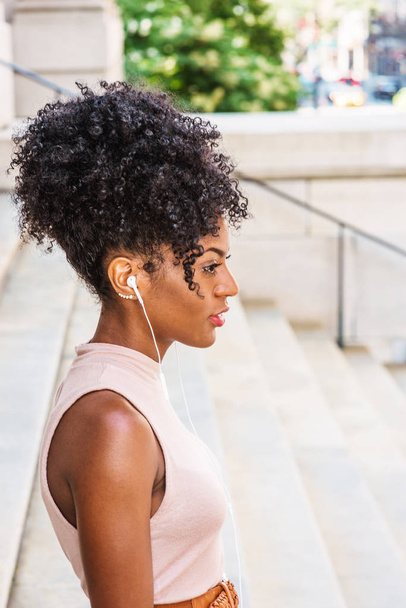Poslech hudby a relaxaci. Mladá africká americká žena s afro účesem na sobě top bez rukávů světle barvy, stojící na schodech mimo kancelářskou budovu, poslechu hudby pomocí sluchátek, myšlení - Fotografie, Obrázek