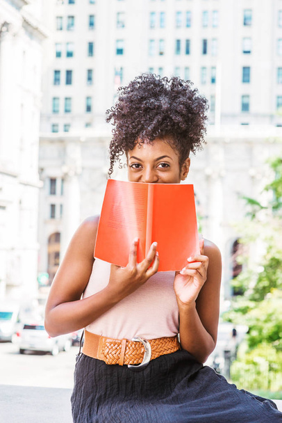 A ler lá fora. Jovem estudante universitária afro-americana com penteado afro, sentada na rua pelo prédio de escritórios de estilo vintage em Nova York, segurando livro vermelho, cobrindo rosto, sorrindo, desfrutando
 - Foto, Imagem