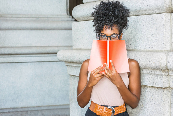 Young African American female college student with afro hairstyle, wearing glasses, standing on street by vintage style column in New York, holding red book, covering face, looking, thinking - Фото, зображення